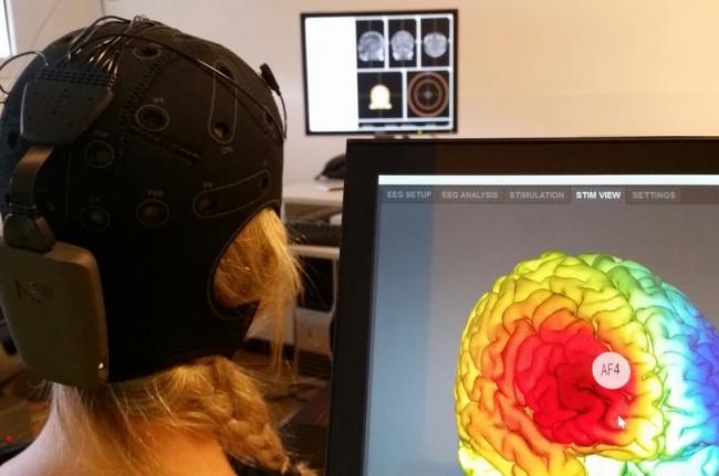 Une personne portant un bonnet en néoprène avec un petit boitier fixé à l'arrière, regardant un écran sur lequel un cerveau en 3D est affiché